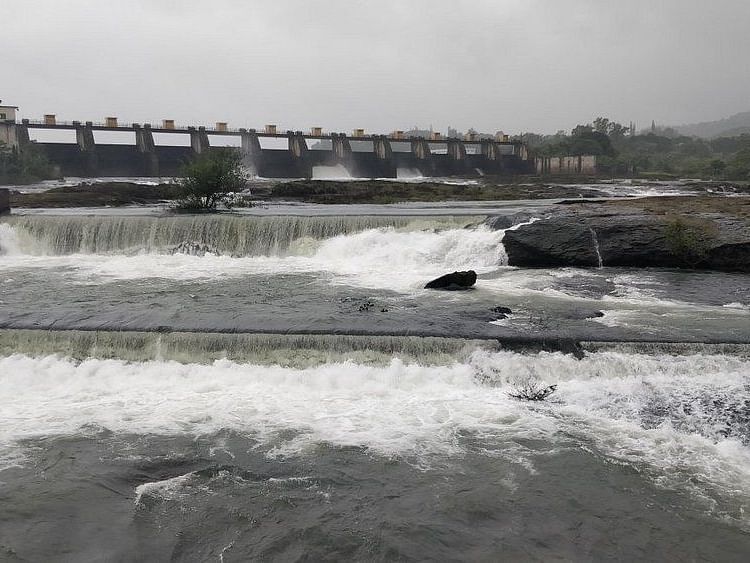 A powerful waterfall cascades over the Khadakwasla Dam, sending a torrent of water downstream. The sky is overcast, and the water appears white and foamy.